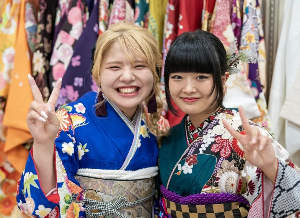 出雲・米子の成人振袖・成人袴のレンタル・明るい青と緑の振袖を着て、店頭で笑顔でポーズをとる2人の女性。豊富な最新トレンドの振袖ラインナップを象徴。